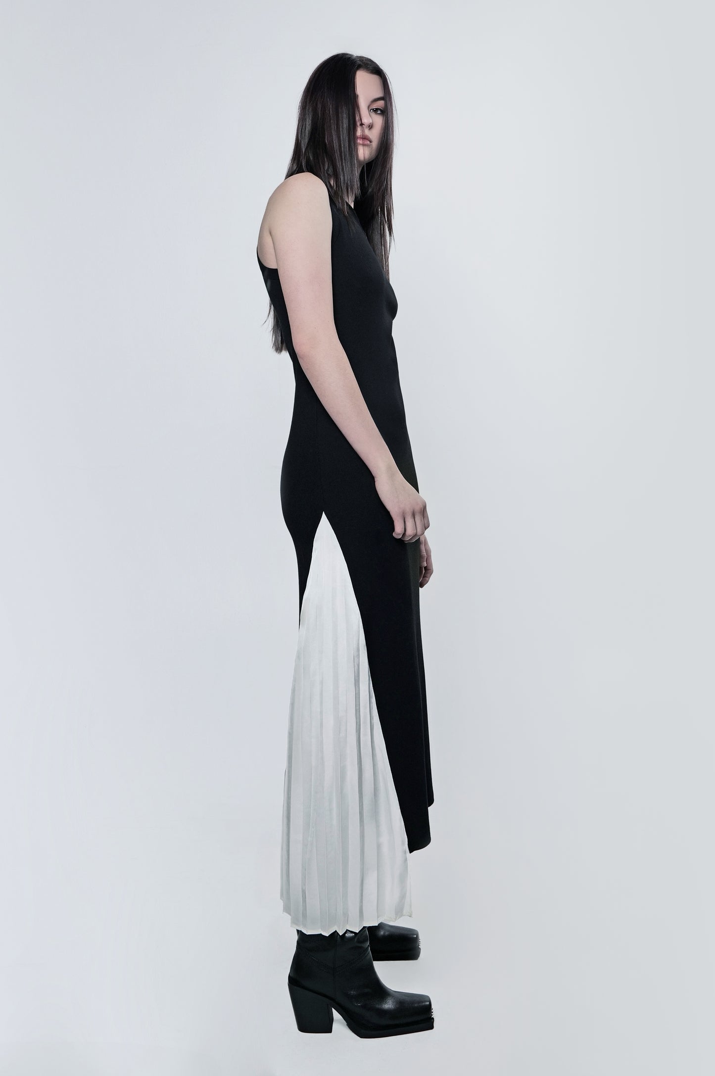 Side view of a woman wearing a black ribbed maxi dress with a fitted silhouette and contrast pleated panel.