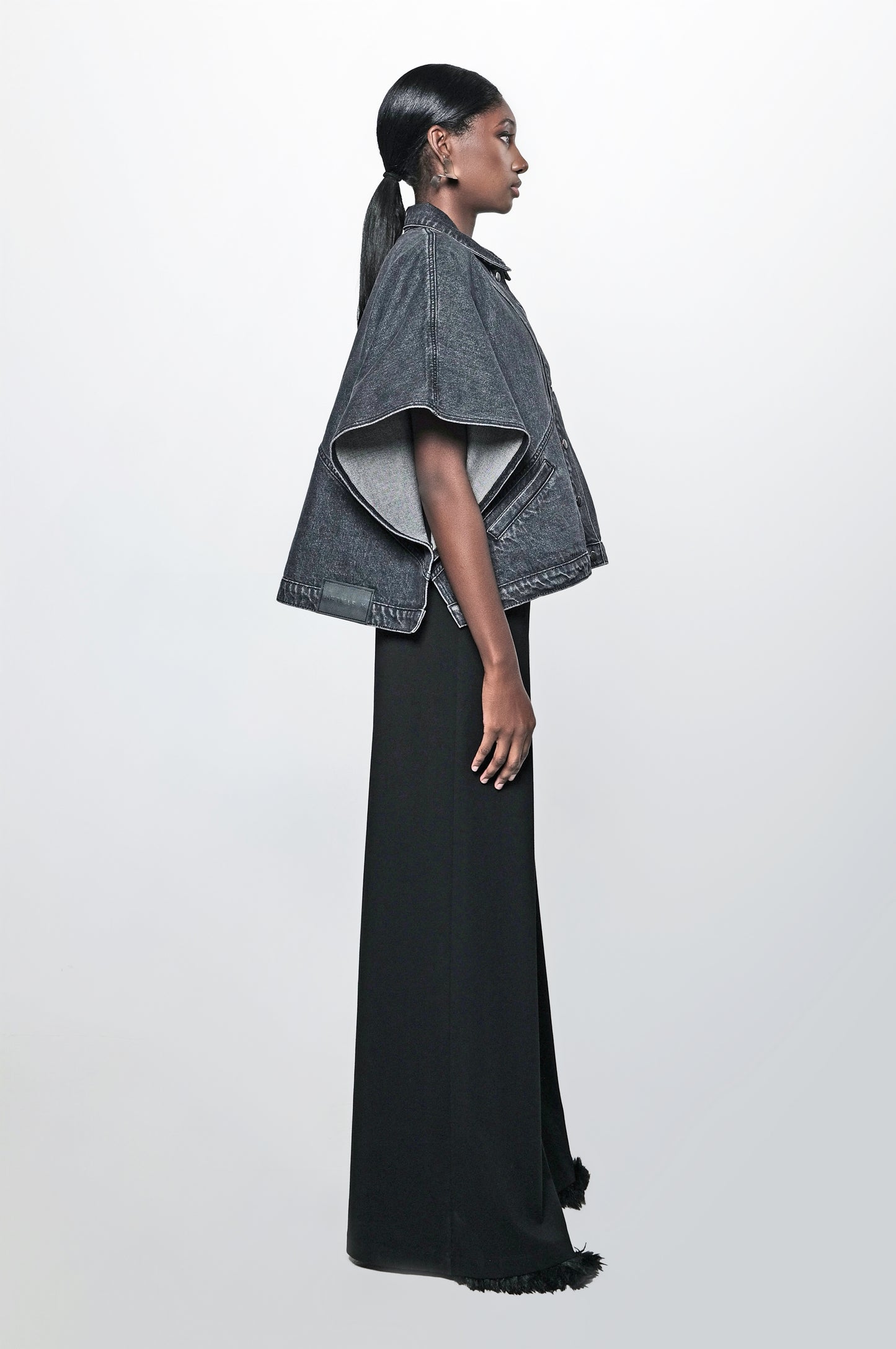 Side view of a woman wearing a black denim cape jacket. The jacket has front pockets and a relaxed fit.