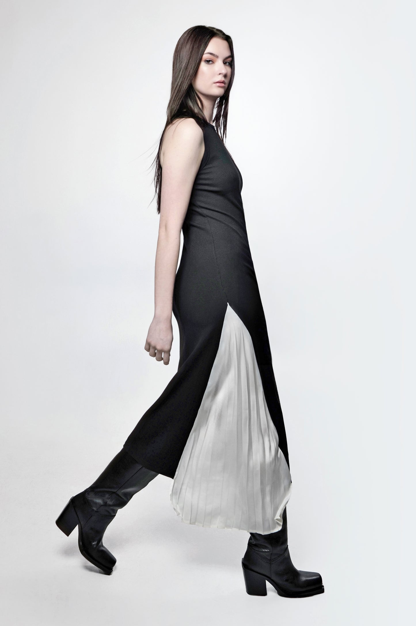 A woman wearing a sleeveless, black maxi dress with a pleated panel and a fitted racerback design. The dress falls to the floor and she is paired with black boots.