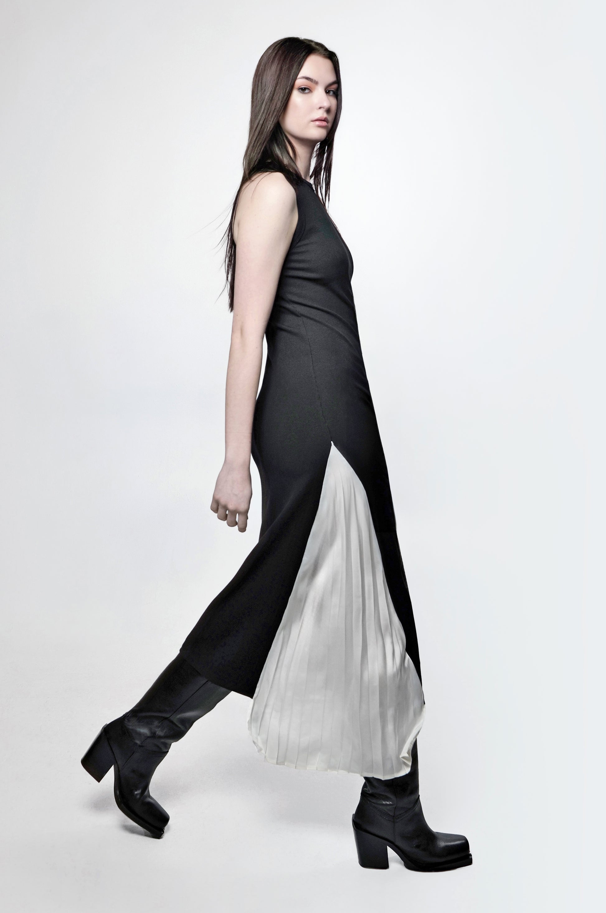 A woman wearing a sleeveless, black maxi dress with a pleated panel and a fitted racerback design. The dress falls to the floor and she is paired with black boots.