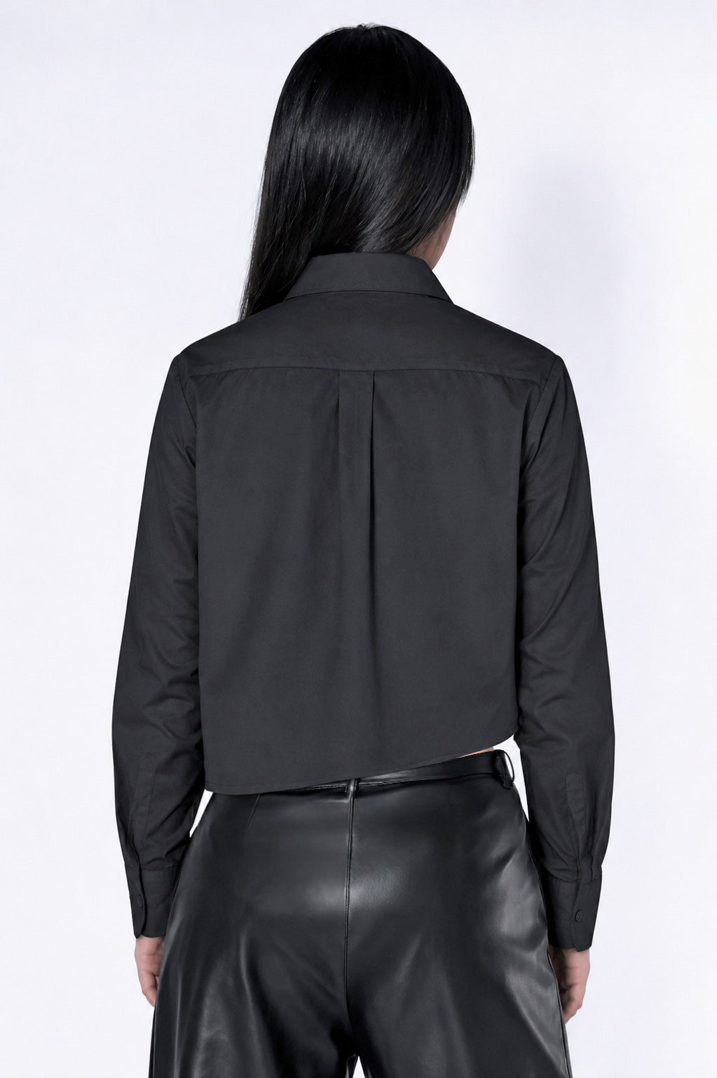 A model shown from the back wearing a black long-sleeve collared shirt with a unique central back pleat detail and black leather pants, against a plain white studio background.