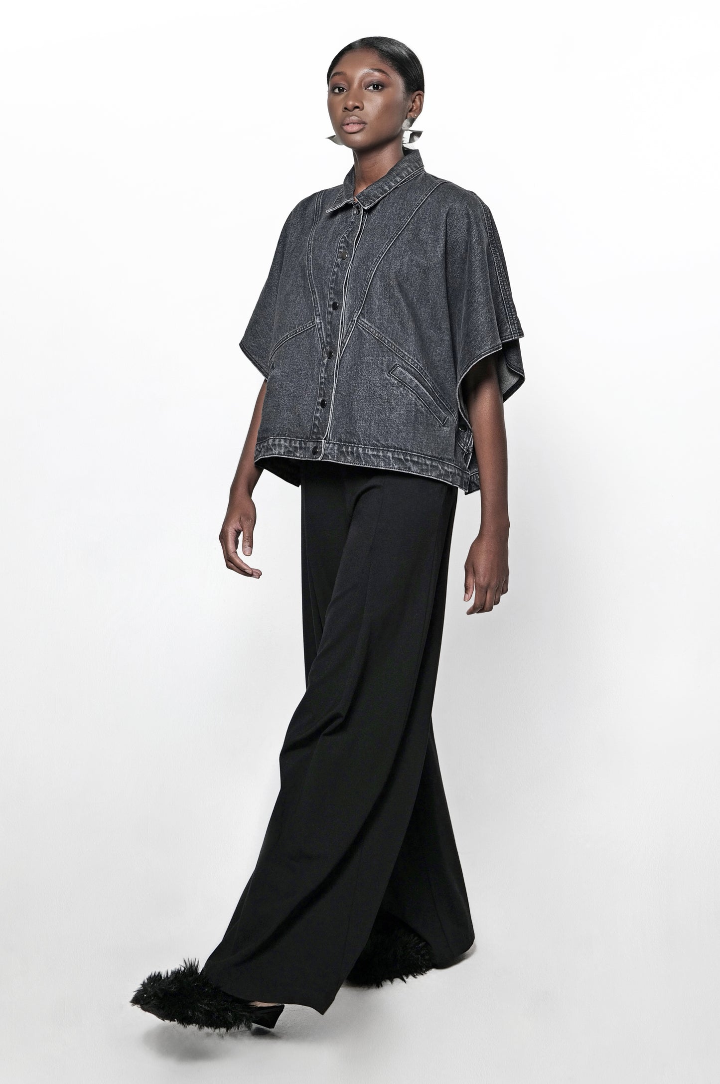 A woman wearing a black denim cape jacket and black pants. The jacket has front pockets and a relaxed fit.
