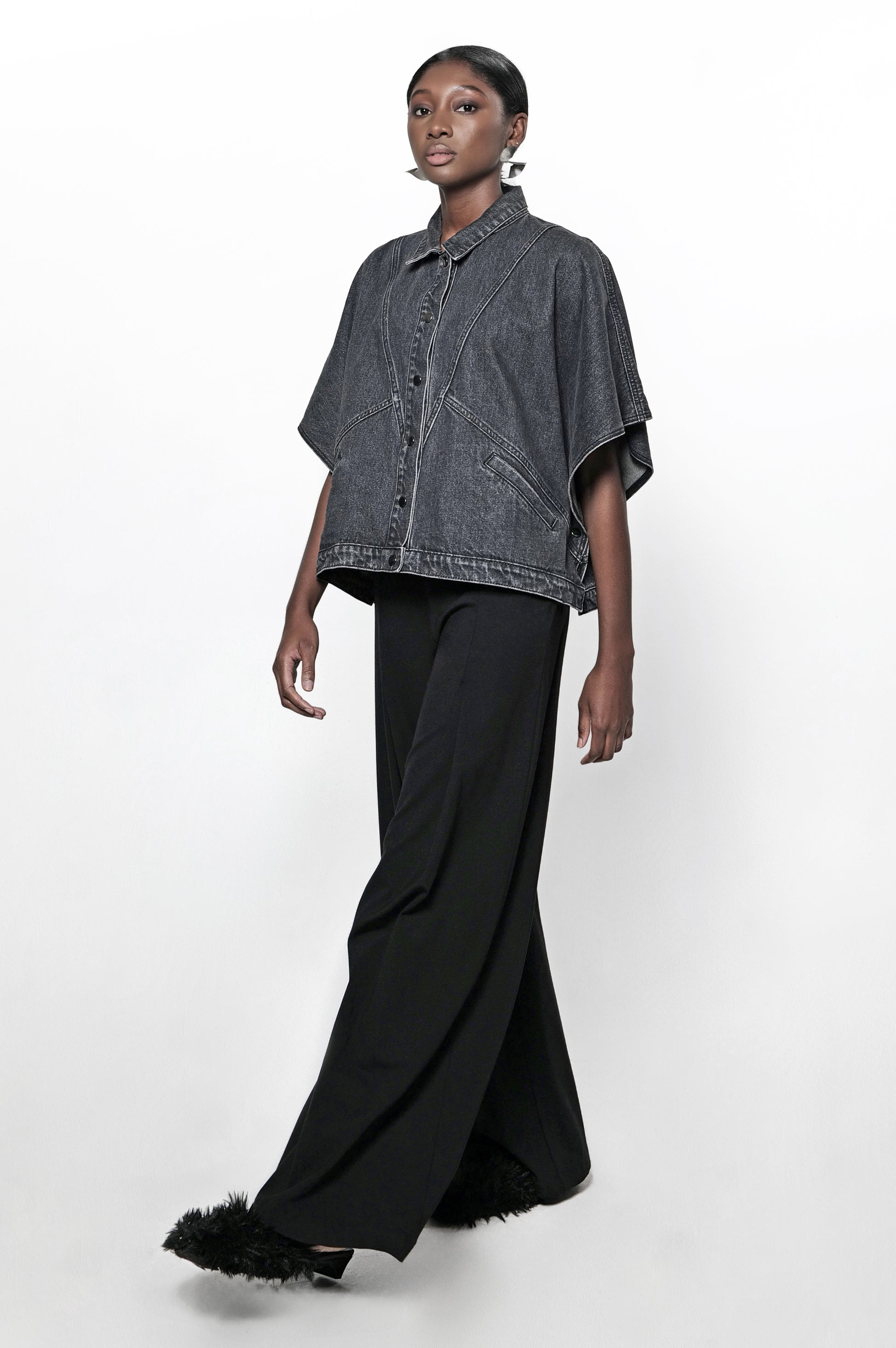 A woman wearing a black denim cape jacket and black pants. The jacket has front pockets and a relaxed fit.