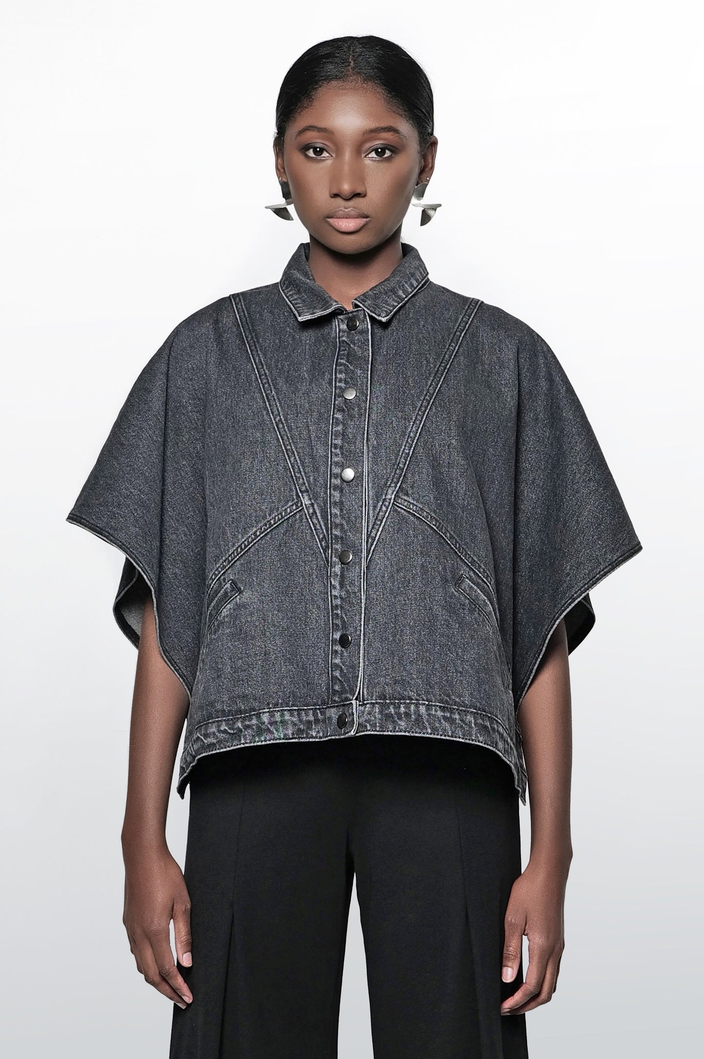A woman wearing a black denim cape jacket with a collar, short sleeves, and snap closures. The jacket has front pockets and a relaxed fit.