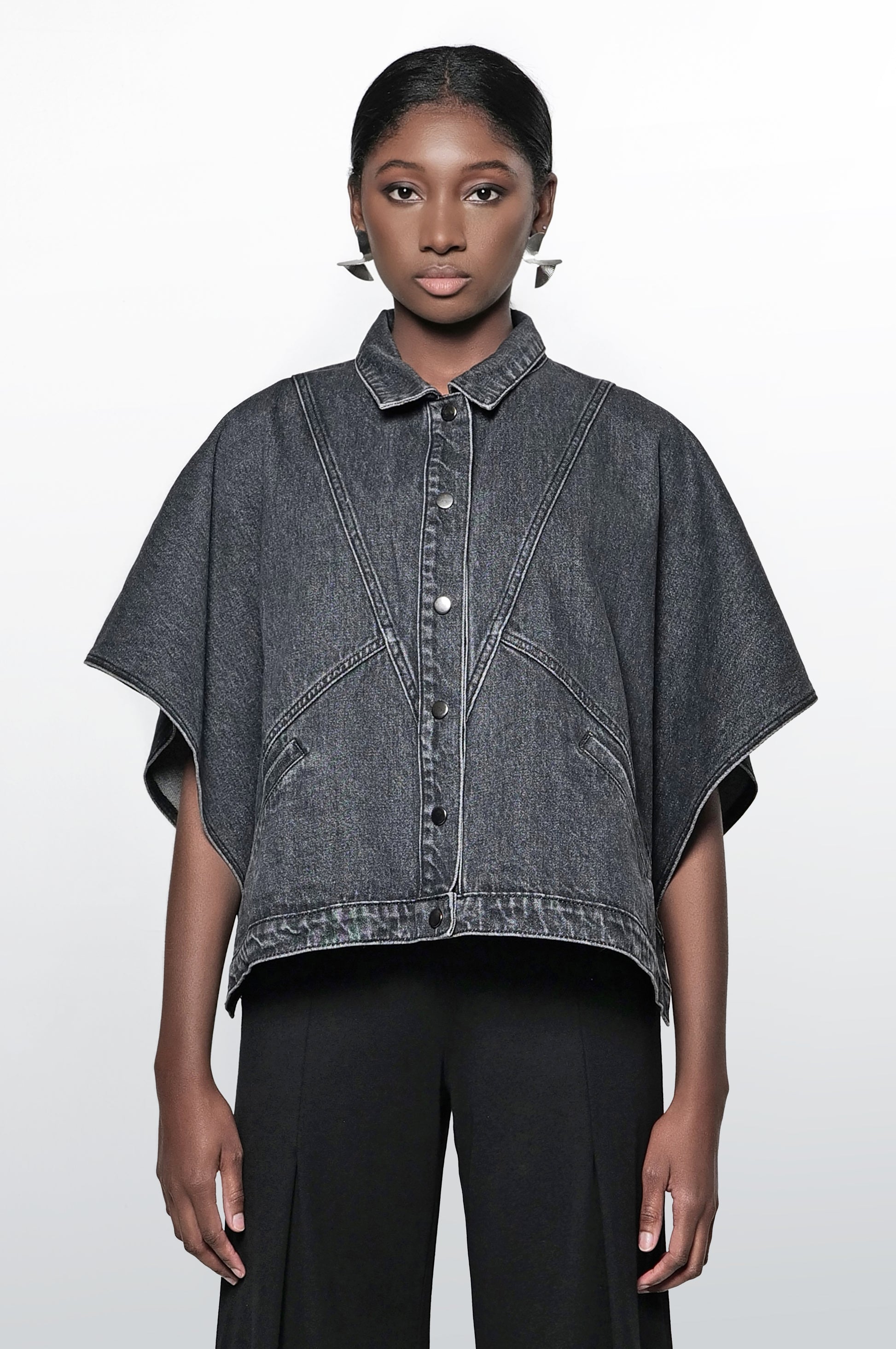 A woman wearing a black denim cape jacket with a collar, short sleeves, and snap closures. The jacket has front pockets and a relaxed fit.