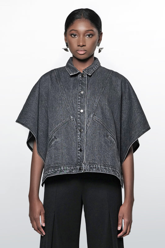 A woman wearing a black denim cape jacket with a collar, short sleeves, and snap closures. The jacket has front pockets and a relaxed fit.