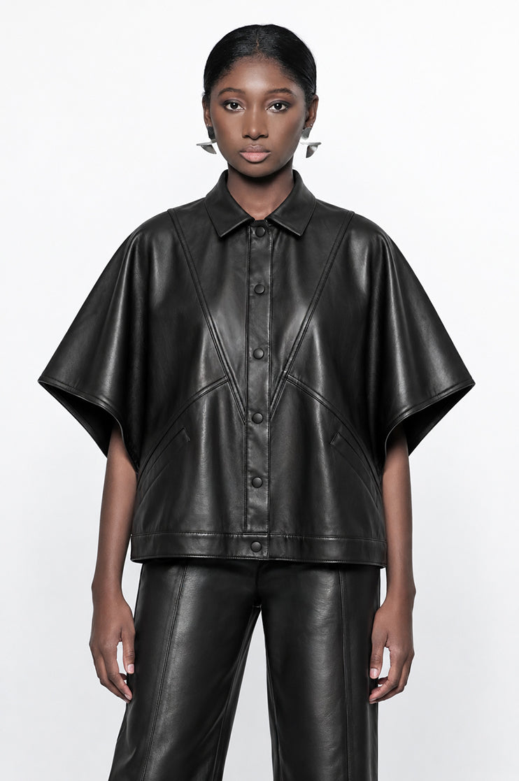 A model wearing a black leather-like short-sleeve button-front top with a collar and matching black leather pants, photographed against a plain light gray studio background.