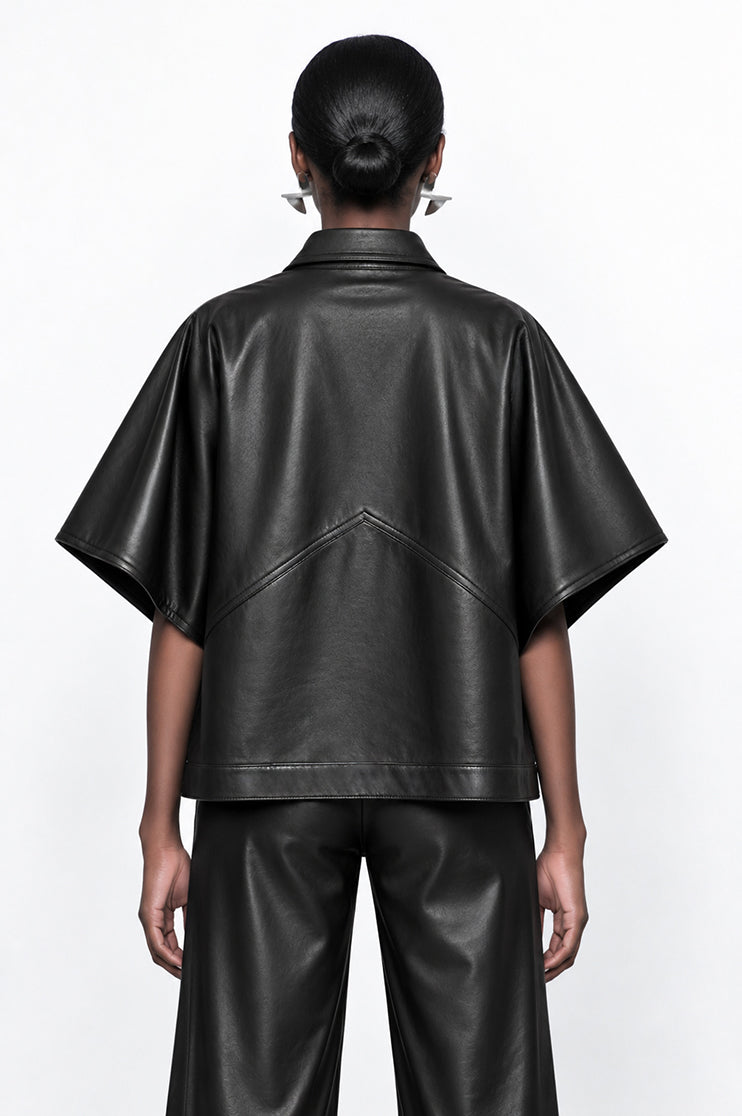 A model shown from the back wearing a black faux-leather short-sleeve cape jacket and matching black wide-leg pants against a plain white background.