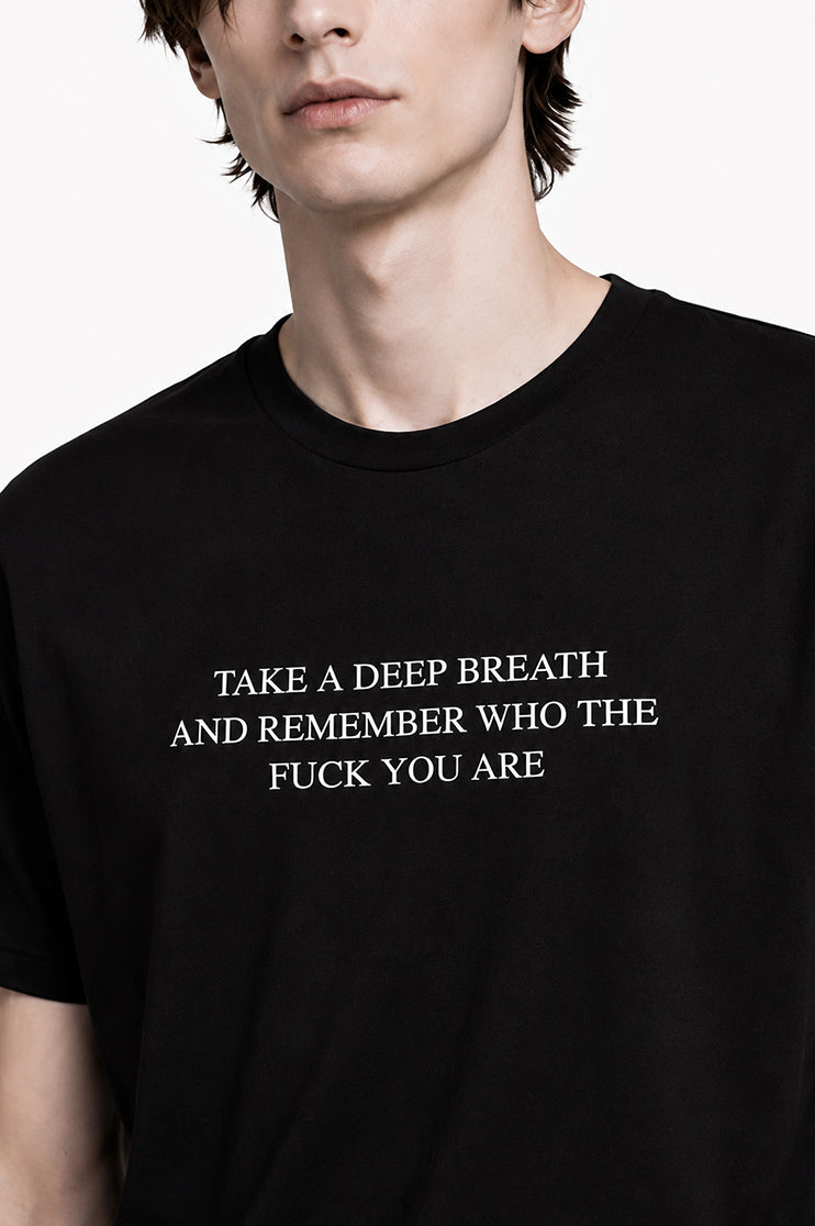 Close up of a man wearing a black t-shirt that says "take a deep breath and remember who the fuck you are"